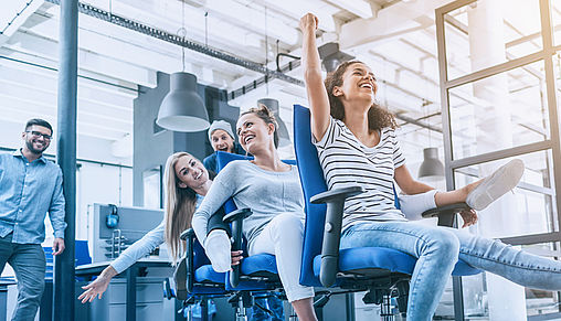 Frauen sitzen hintereinander auf Bürostühlen & halten das Bein der hinteren
