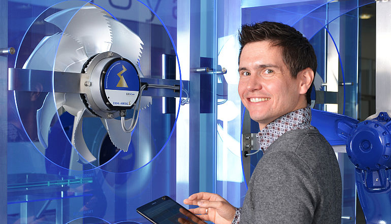 Junger lächelnder Mann steht mit einem Tablet vor einem ZIEHL-ABEGG Ventilator