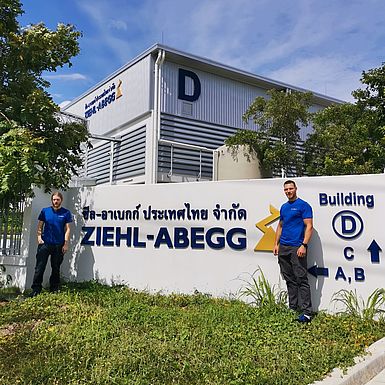 2 Männer  vor dem Standort in Thailand, zwischen ihnen ein  Logo von ZIEHL-ABEGG